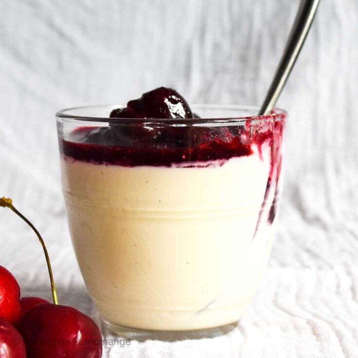 A dessert glass with cherry panna cotta on a white napkin with three whole cherries.