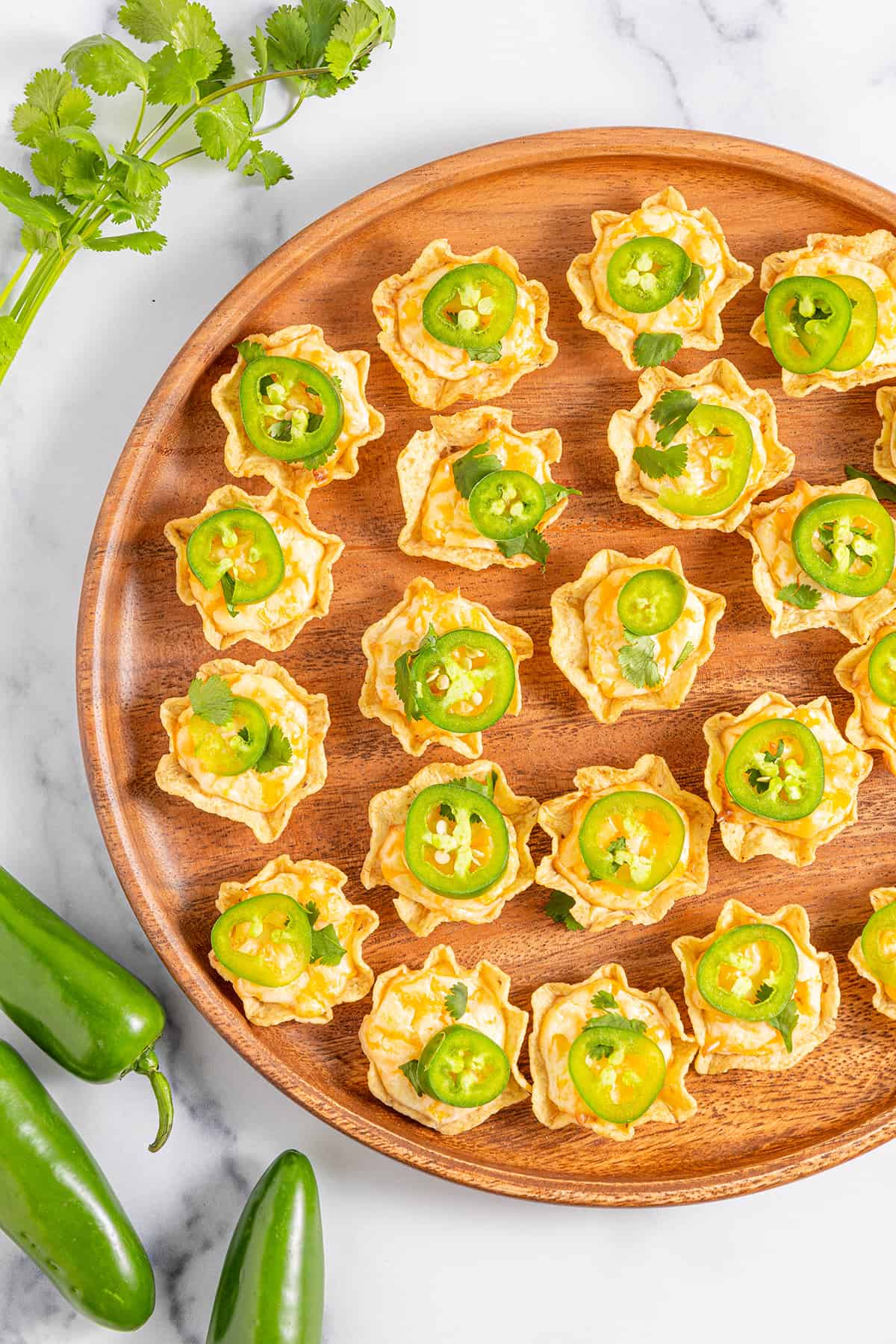 Several cheesy jalapeno bites on a round wooden serving platter with fresh jalapeños and a large sprig of fresh cilantro on a white counter.