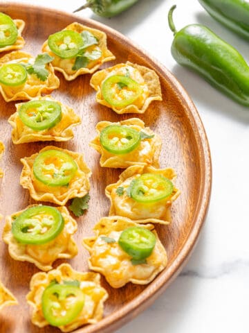 Several Tostitos scoop chips filled with a cheese mixture and topped with fresh sliced jalapeños and cilantro on a round wooden serving tray with three fresh jalapeños on a white counter top.