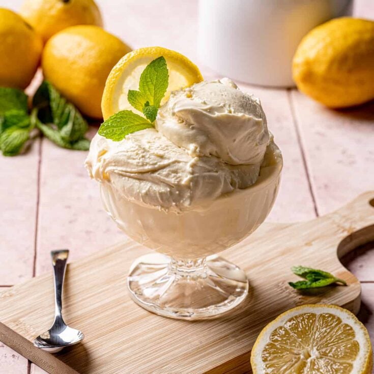 A dessert glass with lemon cream gelato topped with a slice of fresh lemon and fresh mint leaves on a wooden serving board.