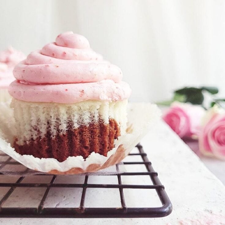 One neapolitan cupcake with strawberry frosting on a black white rack with two pink roses.
