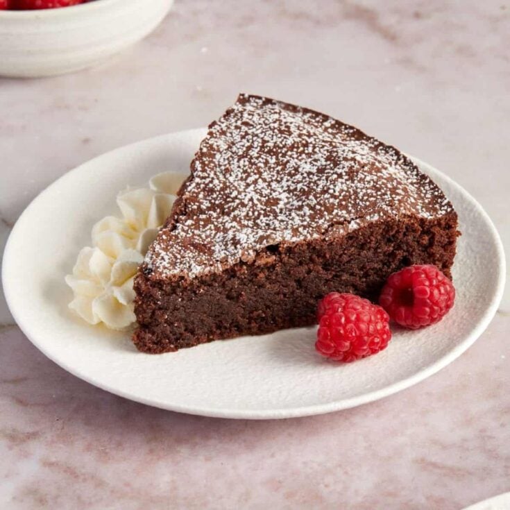 A slice of chocolate flourless cake on a white dessert plate with two fresh raspberries.