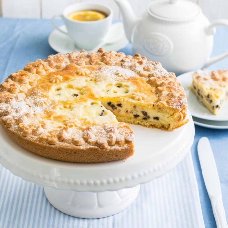 A large ricotta cake on a white cake stand with one slice removed on a serving plate.