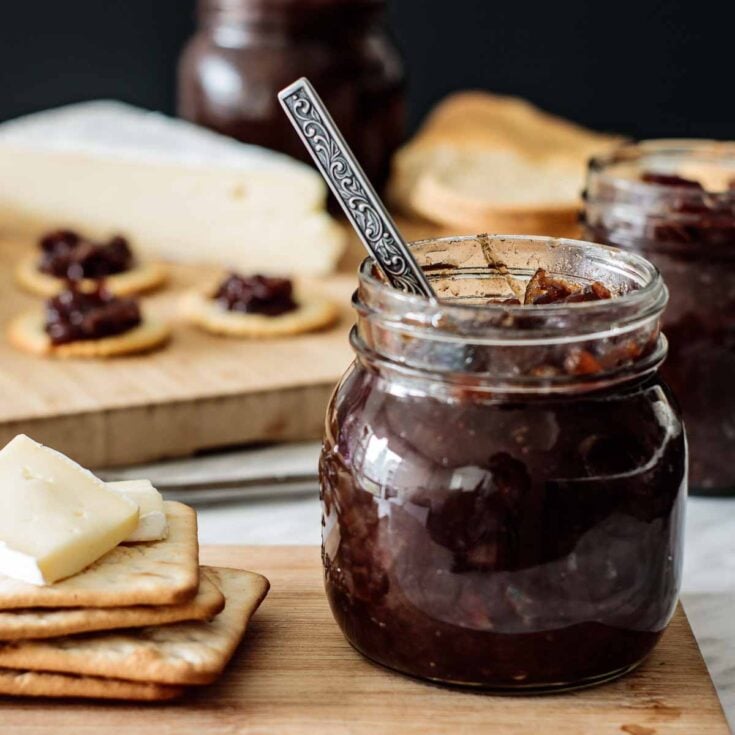 A a silver spoon dipped in to a glass jar with red onion chutney next to some cheese and crackers on a wooden serving platter.