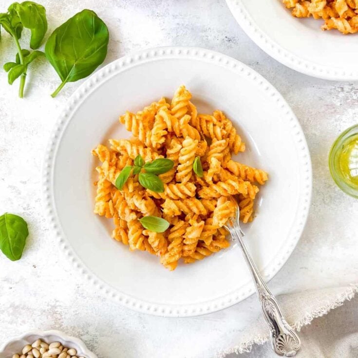 A large white bowl with red pepper pasta topped with fresh basil leaves and silver fork.