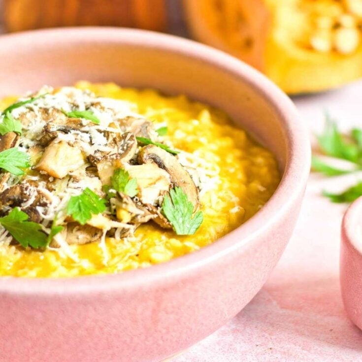 A pink bowl with pumpkin risotto topped with cooked mushrooms, shredded parmesan cheese, and parsley leaves.