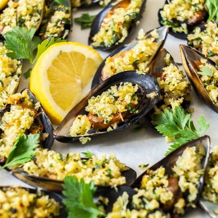 Several baked mussels topped with seasoned bread crumbs garnished with fresh parsley and a wedge of lemon.