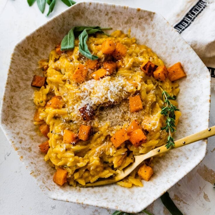 A large bowl with cooked orzo in a creamy orange sauce with diced butternut squash, parmesan cheese and thyme on top.