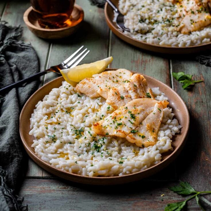 Two pieces of cooked dover sole on lemon risotto in a large serving bowl with a wedge of lemon.