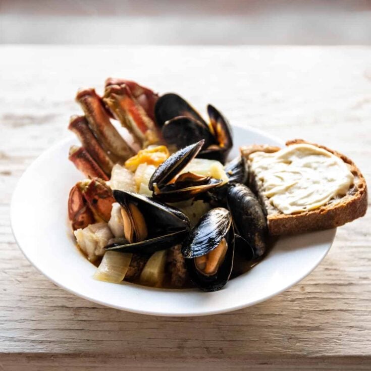 A large white bowl of cioppino soup with clams, crab and white fish and a buttered half slice of bread.