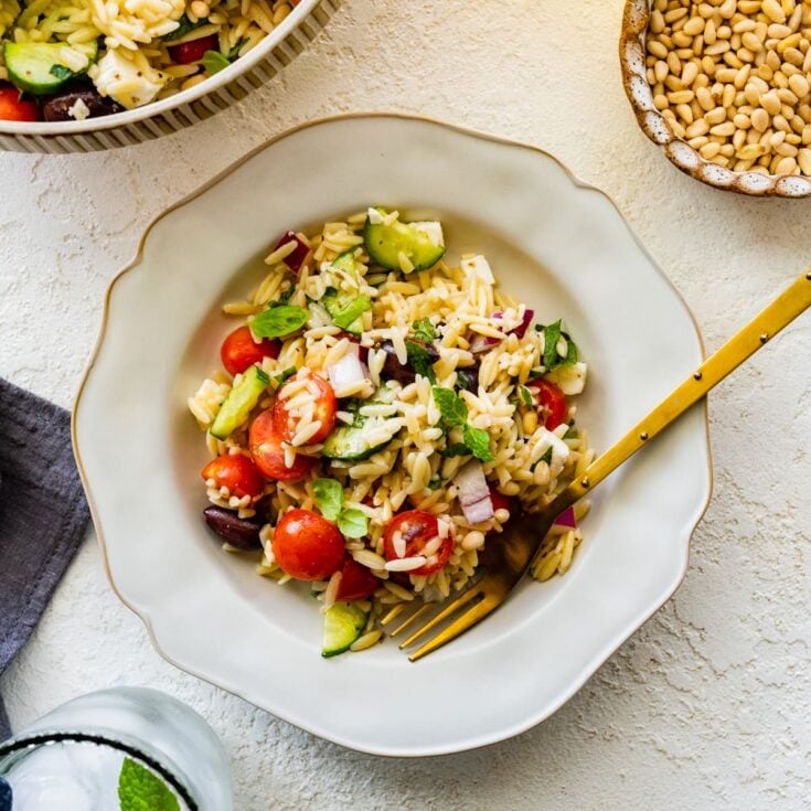 A white serving bowl with halved red cherry tomatoes, sliced cucumbers, Kalamata olives, chopped red onions, orzo and mint leaves.