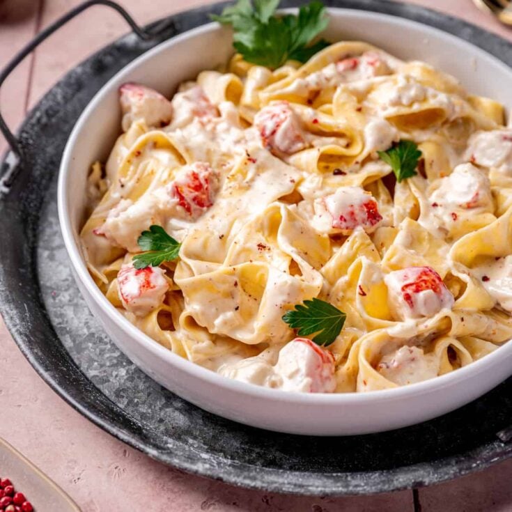 Pappardelle pasta tossed in a champagne cream sauce with large chunks f cooked lobster in a white bowl.
