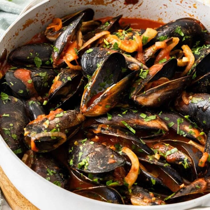Several steamed mussels in a tomato broth topped with fresh chopped parsley in a white serving bowl.