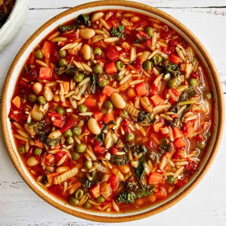 A large bowl with cooked vegetables including peas, carrots, diced tomatoes, spinach, white beans and orzo pasta in a tomato based broth.
