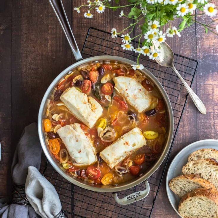 Poached white fish with halved cherry tomatoes, kalamata olives, and garlic in a stainless steel skillet.