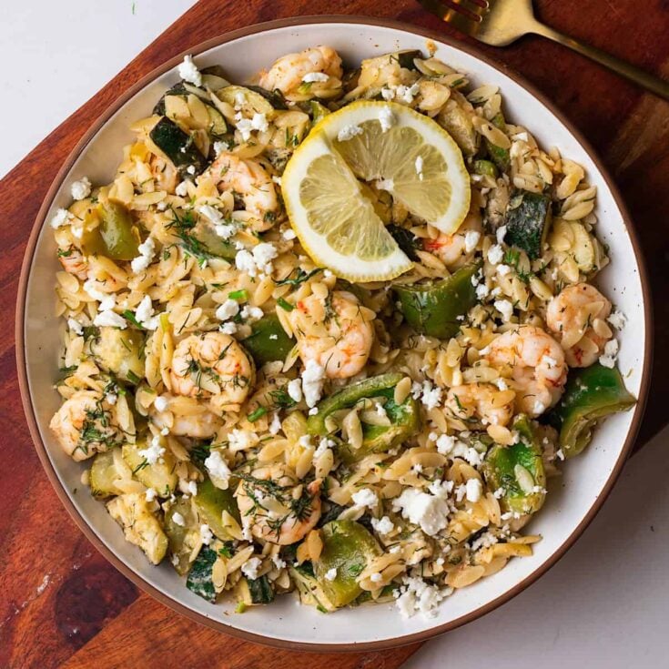 A large white round serving platter with cooked orzo, shrimp, and green bell peppers sprinkled with crumbled feta cheese and dill.
