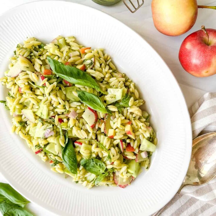 A white serving platter with orzo with pesto, radishes, chopped red onions and fresh basil leaves.